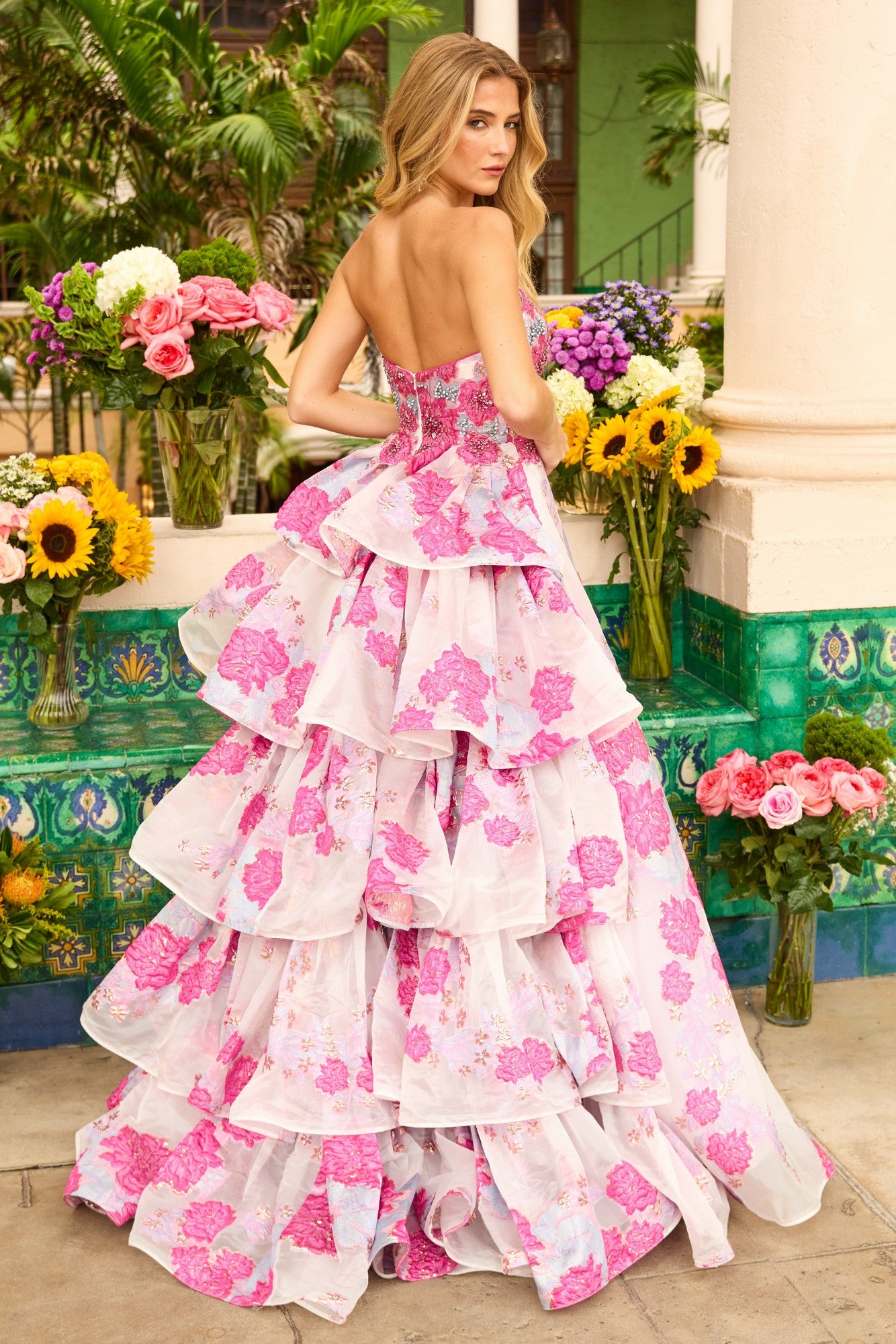 A woman stands outdoors among vibrant flowers and greenery, facing away in the Ava Presley 42292 Long Prom Dress—a strapless floral gown with a tiered ruffle back.